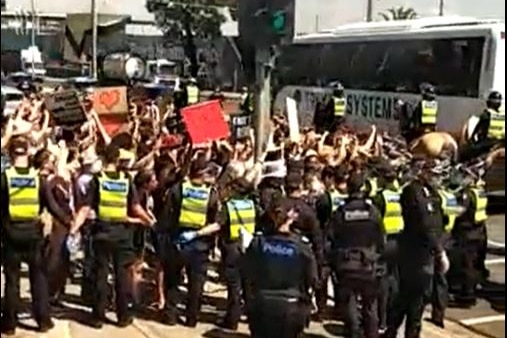 About 15 police officers form a line alongside a large group of protestors, some holding signs