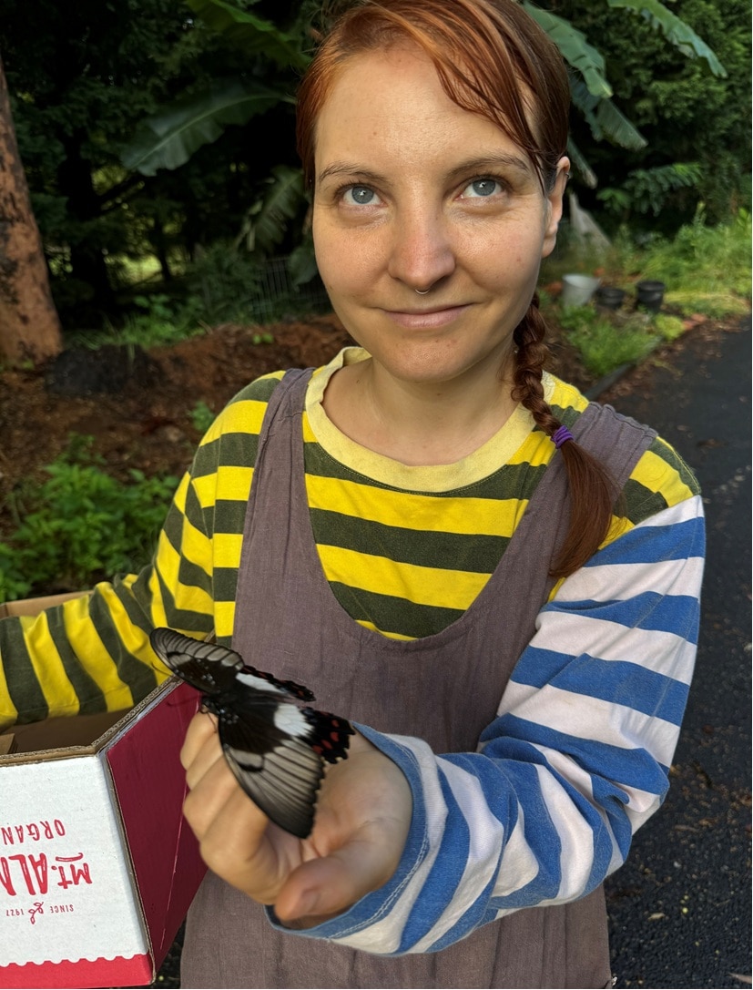 A woman holding a cardboard produce box, with a black and white butterfly resting on her hand.