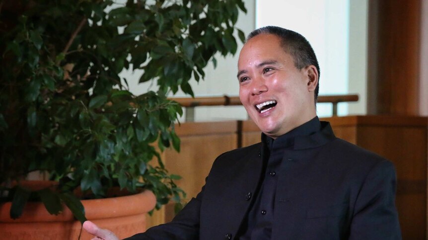 A man in a black suit smiles and gestures with his hands