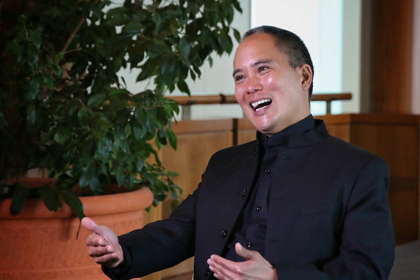A man in a black suit smiles and gestures with his hands
