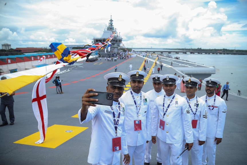 Six navy sailors pose for a selfie on the deck while wearing white dress uniform.