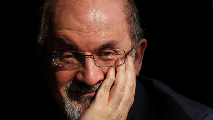 Salman Rushdie holding his left hand up to his face looking at the camera and smiling wearing glasses