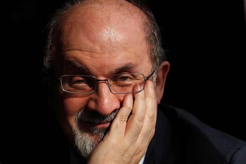 Salman Rushdie holding his left hand up to his face looking at the camera and smiling wearing glasses