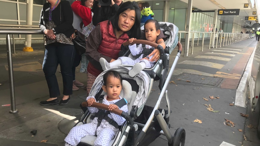 Twin girls sit in a double pram hugged by their mother.