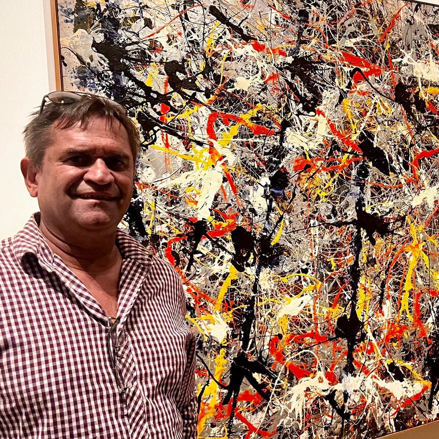 An Indigenous man stands next to an abstract, expressionist painting in the style of Jackson Pollock. 