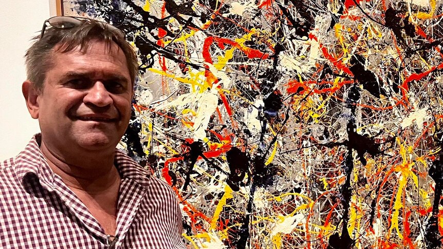 An Indigenous man stands next to an abstract, expressionist painting in the style of Jackson Pollock. 
