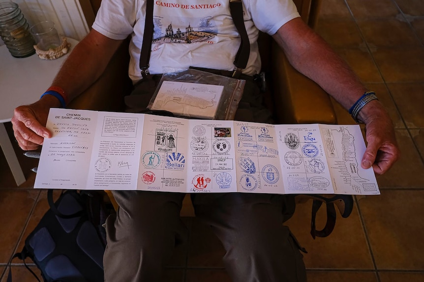 A man wearing suspenders over pants and a white shirt holds out a long pamphelet displaying multicoloured stamps 