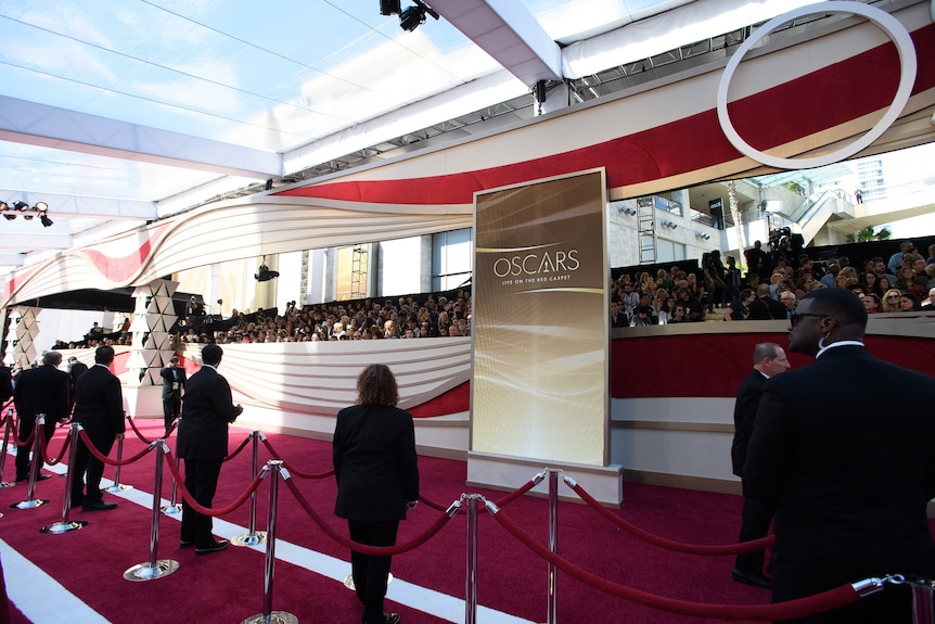 A line of security guards dressed in tuxedos stand on red carpet in front of velvet ropes and people in a grandstand.