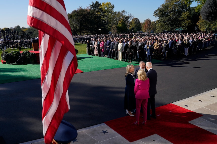 The prime minister, president and their partners speak while a crowd watches on. An American flag is in the foreground.