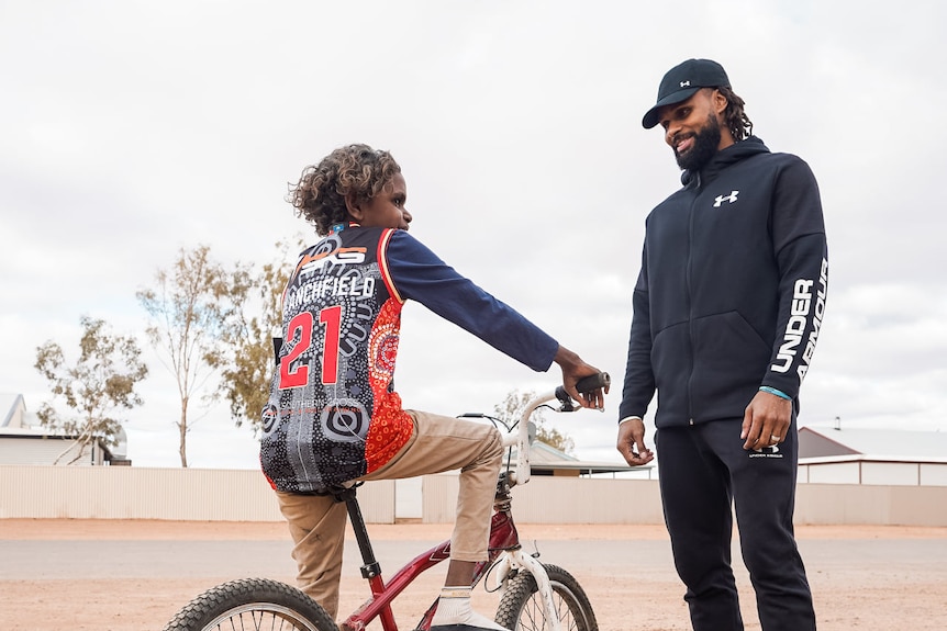 Patty Mills stands looking at a young boy sitting on a bike