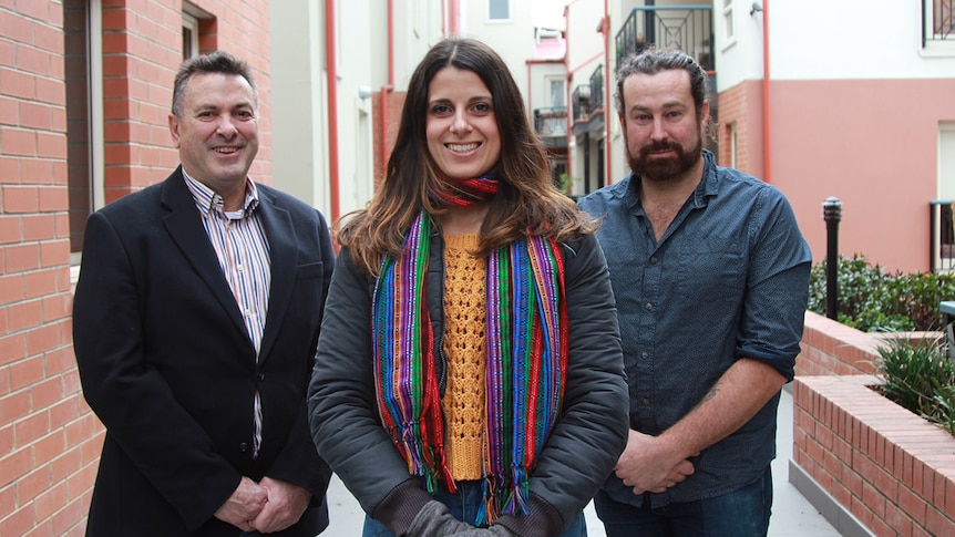 A woman stands between two men in the narrow courtyard of an apartment building.