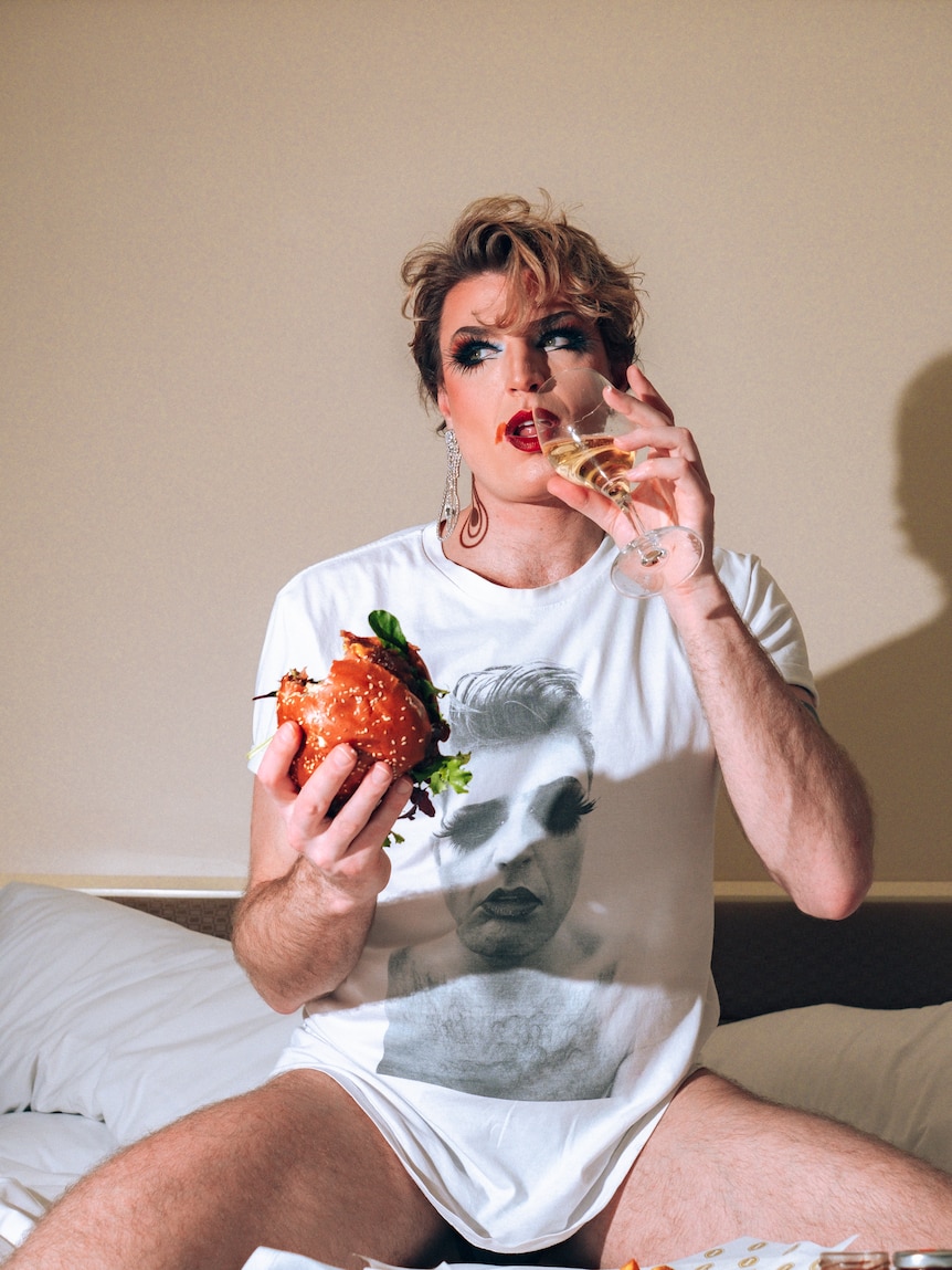 A man in his 30s in smudged drag makeup, in a tshirt with his own face on it, sits in bed with glass of bubbly and a burger