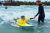 A young girl high high-fives a man in a wet suit as he prepares to stand up on a surfboard