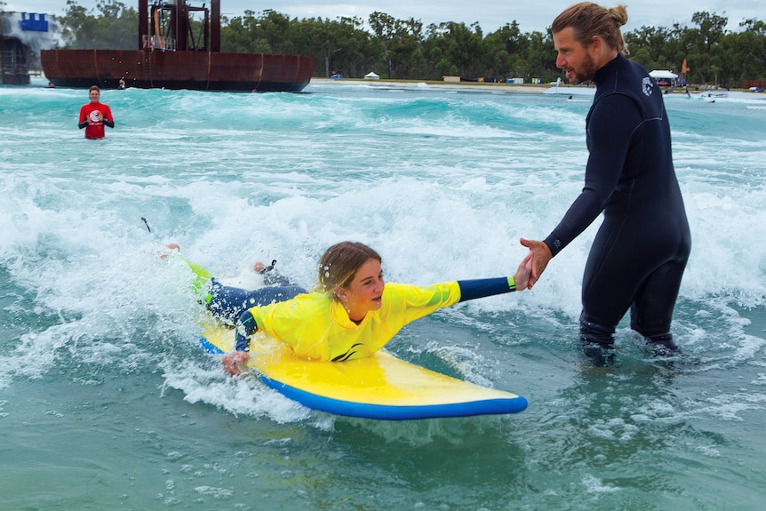 A young girl high high-fives a man in a wet suit as he prepares to stand up on a surfboard