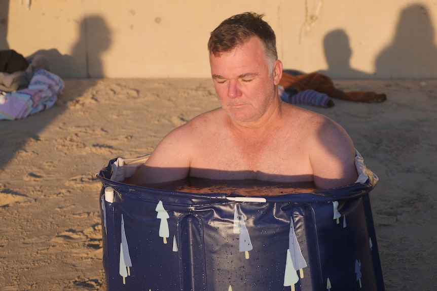 Man sits in small blue bath of icey water with his eyes closed 