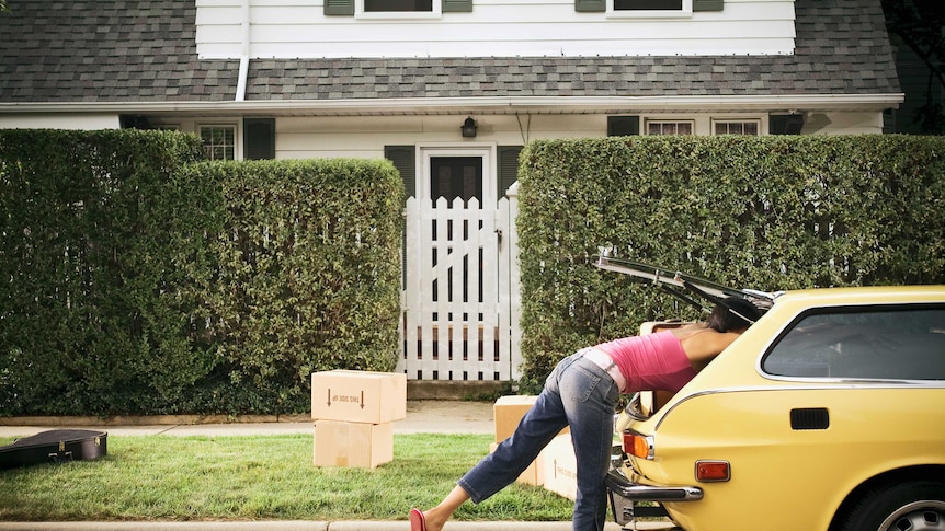 A woman moving house leans into a car boot, surrounded by packing boxes.