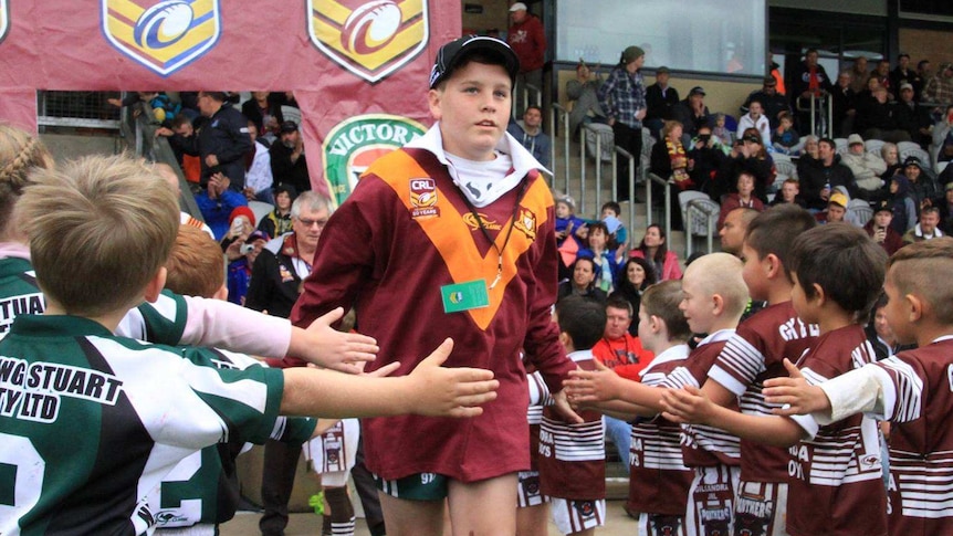 A boy in a footy jersey walks onto a football field through two lines of young players at a stadium