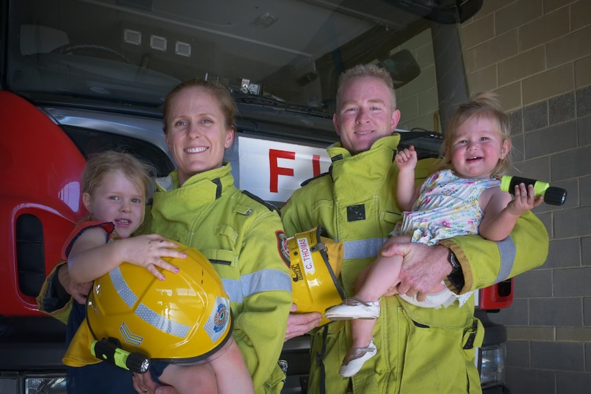 A man and a woman in fire-fighting high-vis uniforms hold a young girl in each of their arms, together inside a fire station