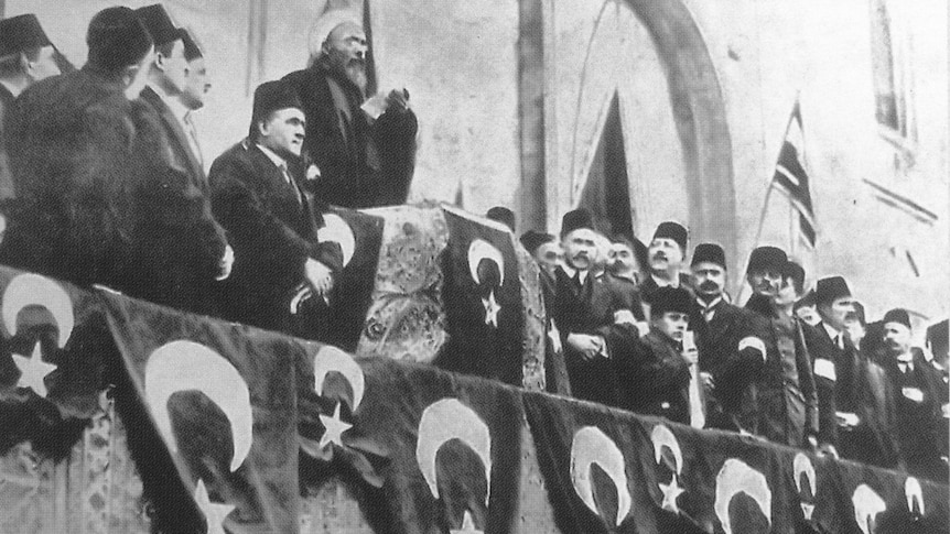 A Muslim religious leader talks on a podium with other men standing around him