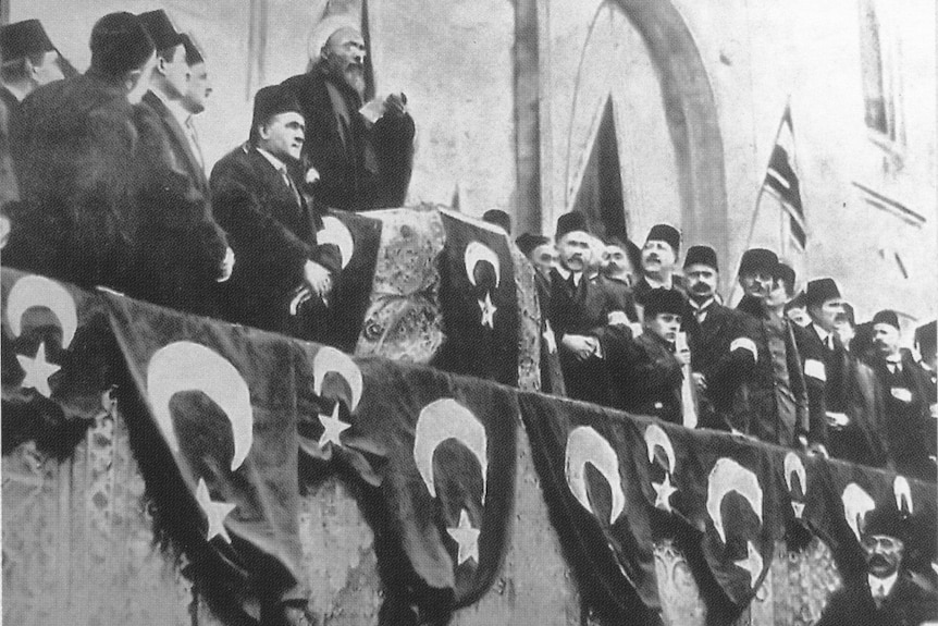 A Muslim religious leader talks on a podium with other men standing around him