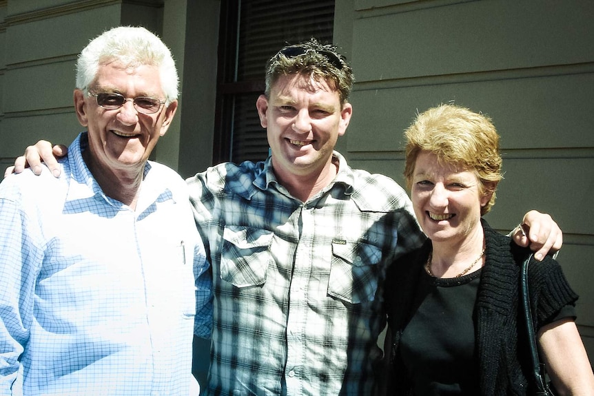 A younger man (centre) stands with arms around an older man and woman who stand either side.