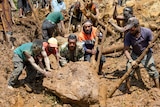 Men use long sticks to lift a large boulder in thick, wet mud looking strained