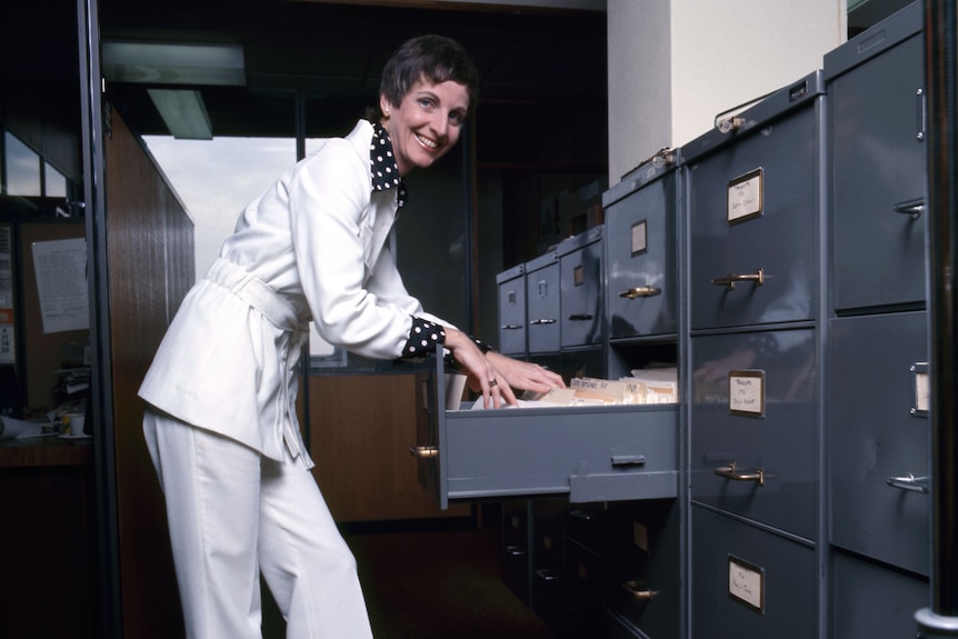 A 1970s photograph of Caroline Jones in white pantsuit at filing cabinet