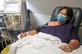 Lea Russ lying in a hospital bed wearing a blue surgical mask, with her arm hooked up to an IV.