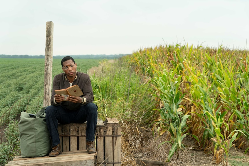 Actor Jonathan Major sitting on a wooden pallet reading a book surrounded by fields in 50s America in TV show Lovecraft Country
