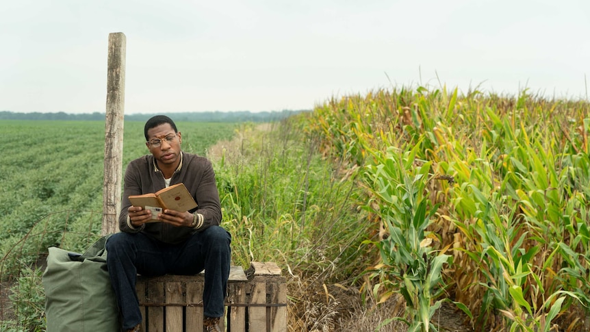 Actor Jonathan Major sitting on a wooden pallet reading a book surrounded by fields in 50s America in TV show Lovecraft Country
