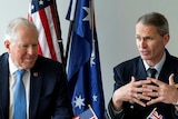 Two men in suits with military pins speak at a desk adorned with small United States and Australian flags