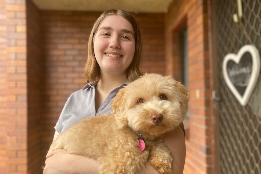 A golden coloured small dog being held by a young woman in her 20s with neck length sandy coloured hair