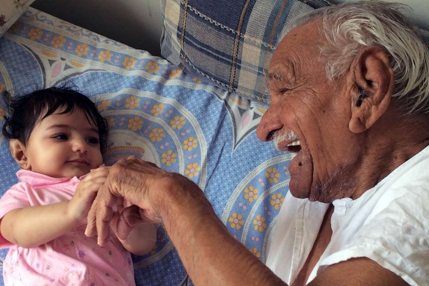 An elderly man and his great-granddaughter play together on a bed.
