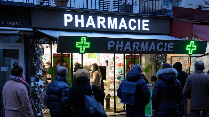 people in winter clothes stand outside a pharmacy at night in Paris