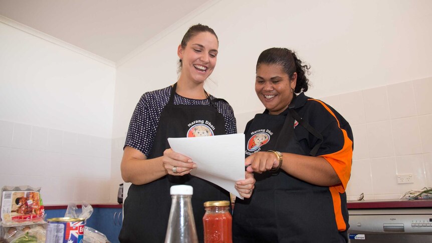 Two women in aprons saying Marang Dhali pointing at a sheet of paper