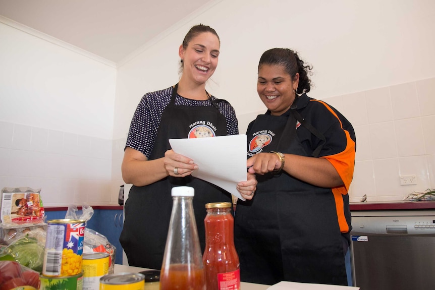 Two women in aprons saying Marang Dhali pointing at a sheet of paper