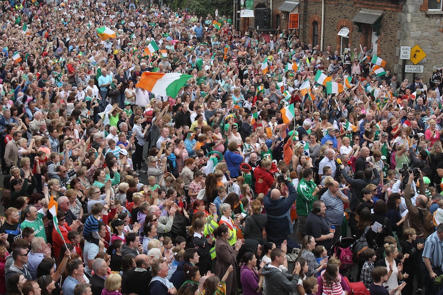 A large crowd of people with Irish tricolour flags stand in a street