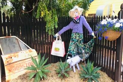 A scarecrow wearing a purple cardigan holds a shopping bag in a garden