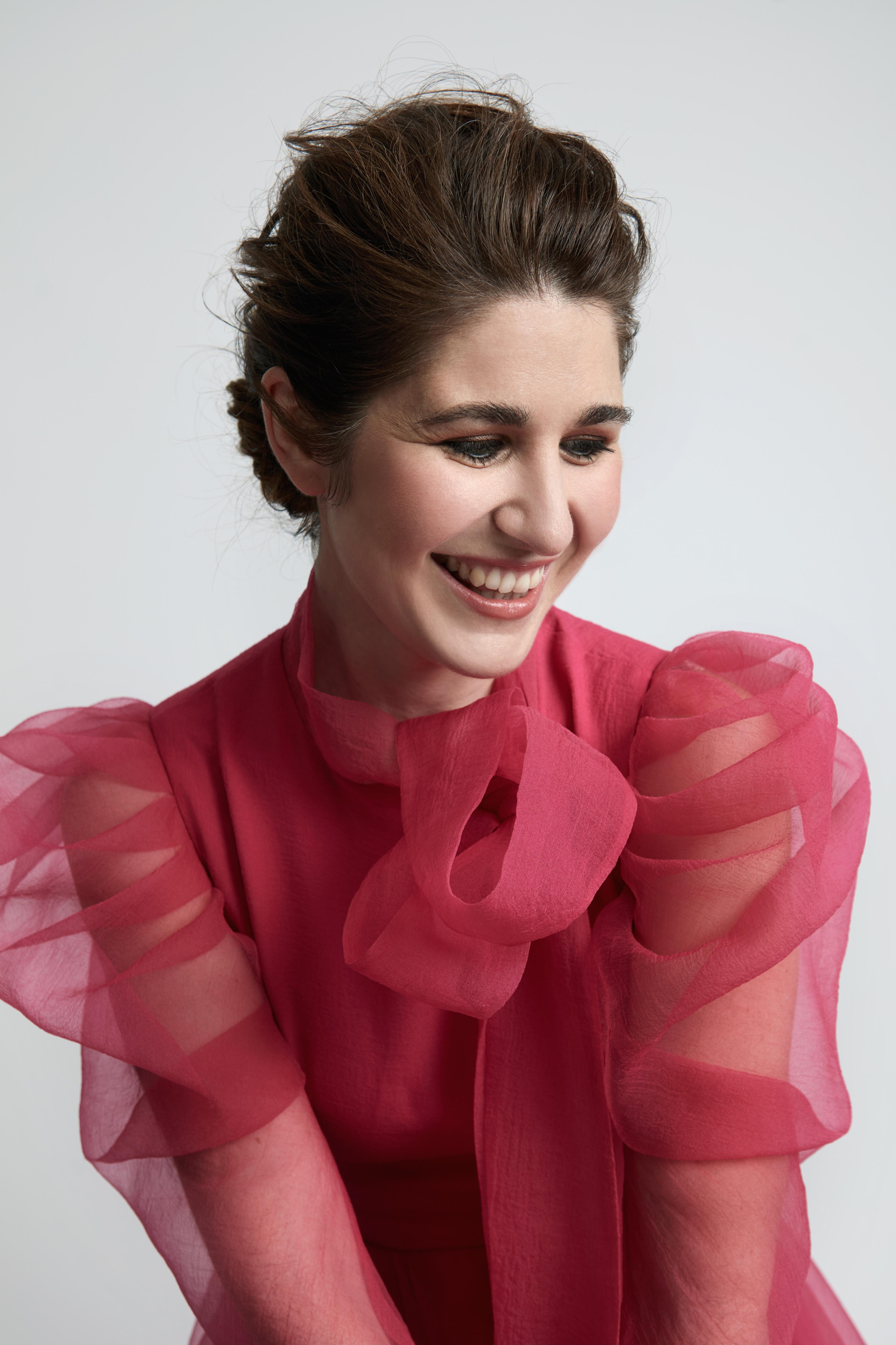 A woman in a sheer red blouse smiles looking toward the ground.