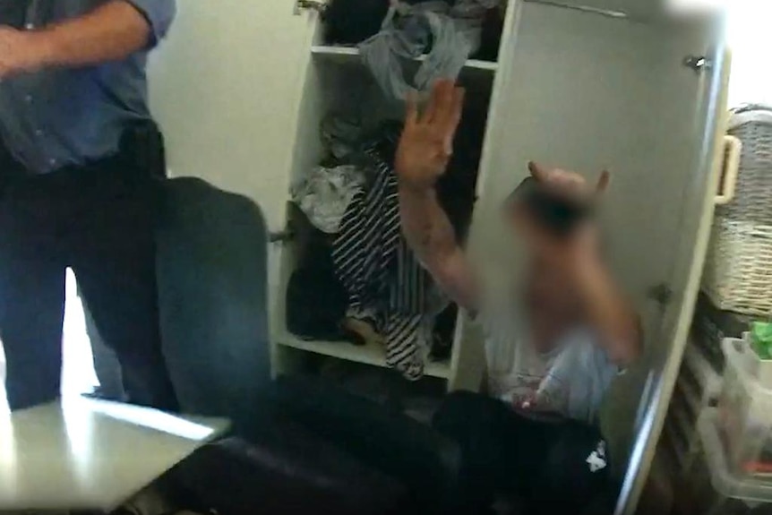 Man squatting in a wardrobe with his hand up.