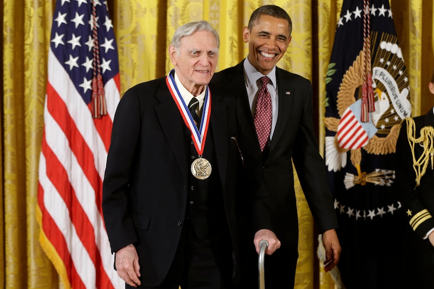 An elderly white man in a suit wears a medal around his neck. Barack Obama stands next to him, smiling. US flags in background.