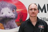 balding man wearing black shirt standing in front of signage with large image of emu.