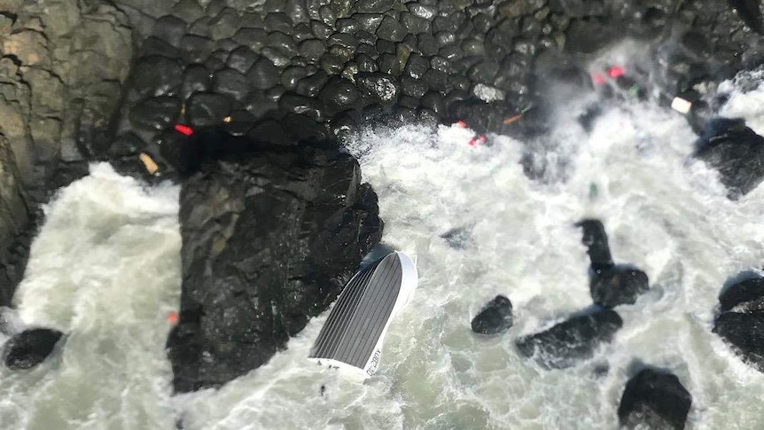 The rough sea smashes against rocks at a cliff base, where a capsized tinny floats