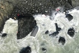 The rough sea smashes against rocks at a cliff base, where a capsized tinny floats