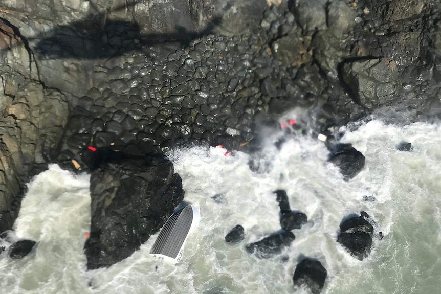 The rough sea smashes against rocks at a cliff base, where a capsized tinny floats