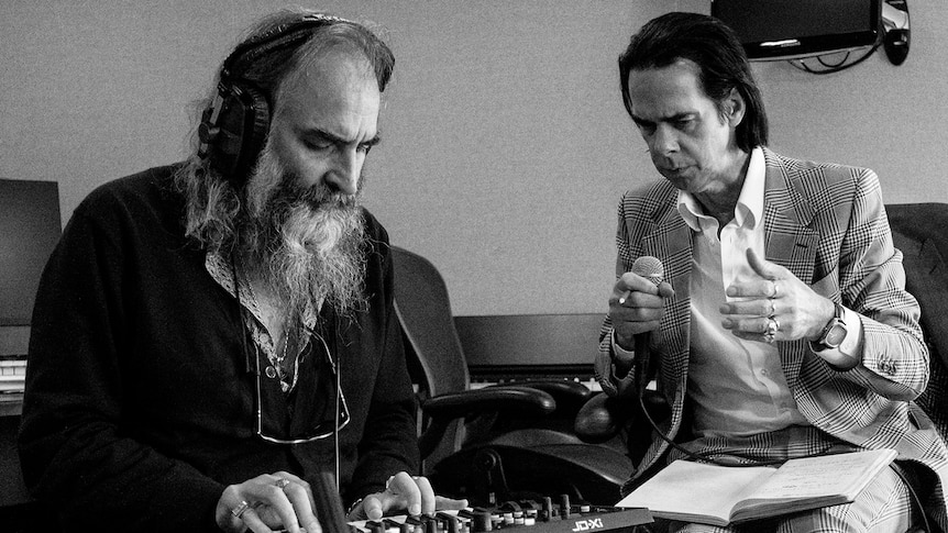 Nick Cave holds a cigarette and microphone, while Warren Ellis plays a synthesizer while wearing headphones
