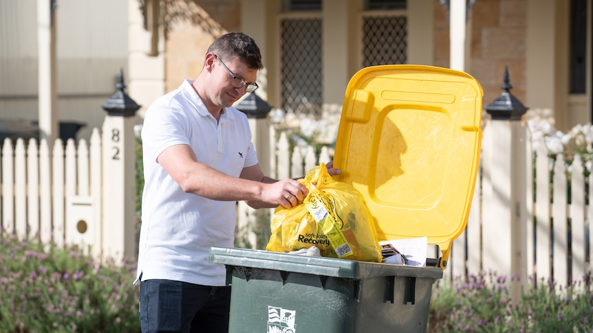Man with white polo shirt putting yellow plastic bag into yellow-lidded green rubbish bin in front of house with white fence