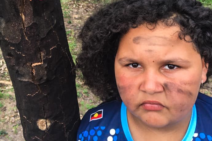 Young girl with charcoal on her face standing next to burnt tree with epicormic shoots on trunk