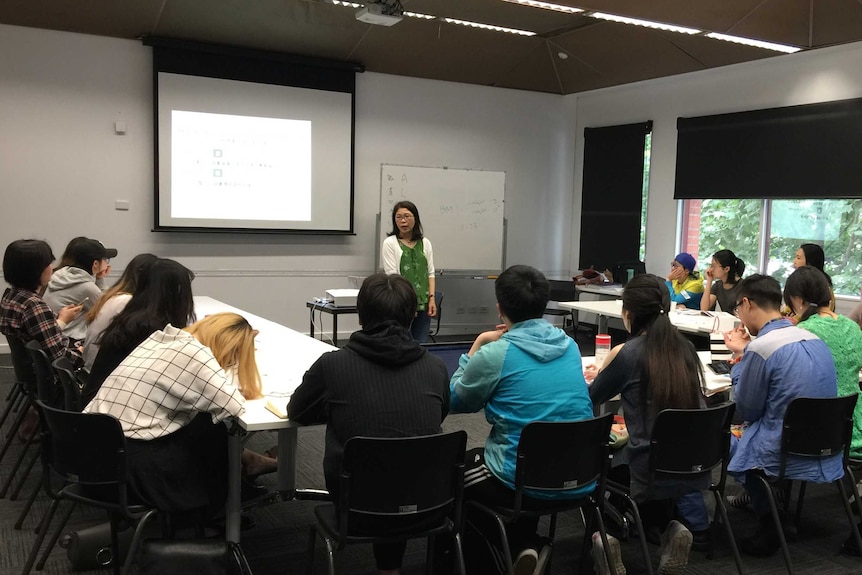 A woman stands in front of a classroom of Chinese-speaking students with a screen and projector behind her.