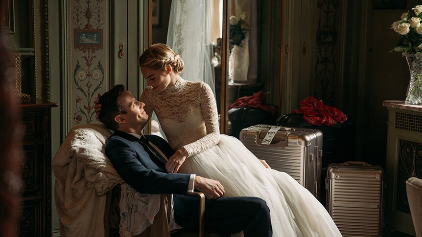 Samara Weaving in lace wedding dress sits on the lap of Mark O'Brien in suit, both look lovingly towards each other in room.